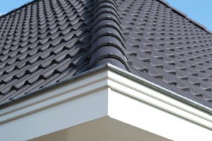 Detail van een zwart zadeldak met dakpannen en witte dakrand. Zicht op een schoorsteen tegen een heldere blauwe lucht.